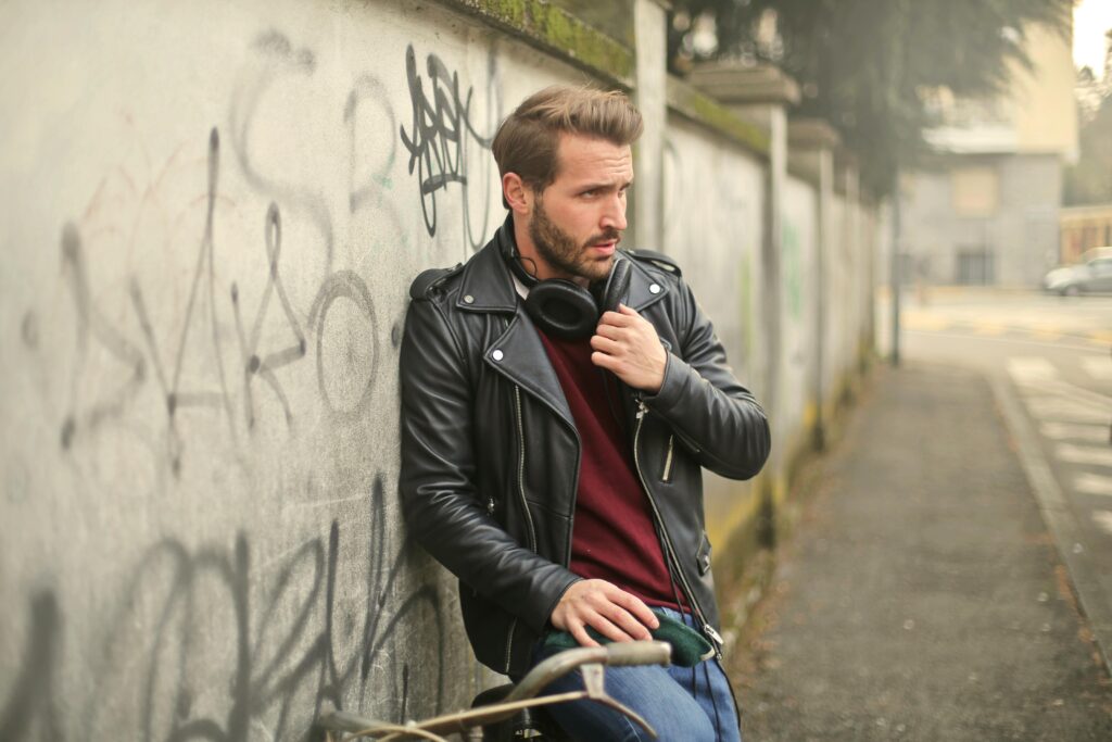 Fashionable man in a black leather jacket with headphones, leaning against a graffiti wall outdoors.